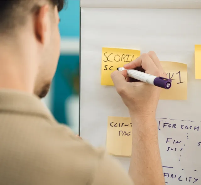 A man writing on a post it note stuck to a whiteboard using a marker.