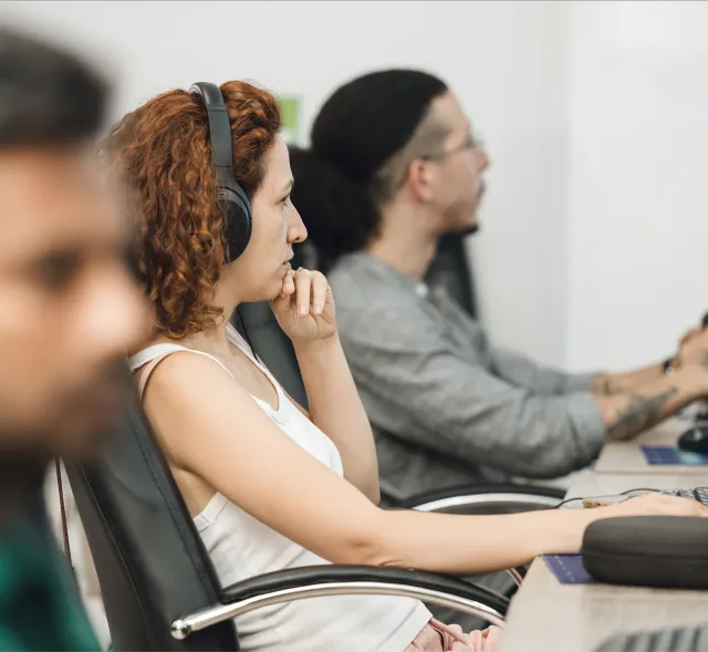 A focused group in an office setting. Foreground: woman with red hair and headphones, deep in thought. Background: blurred people working on computers.