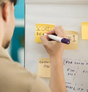A man writing on a post it note stuck to a whiteboard using a marker.
