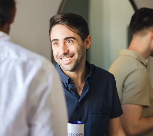 A man in a navy shirt smiles while holding a mug, engaging in conversation with another person.