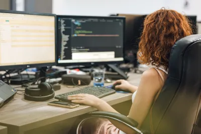 A woman working on her computer using dual monitors, the left displaying a website and the right displaying code.
